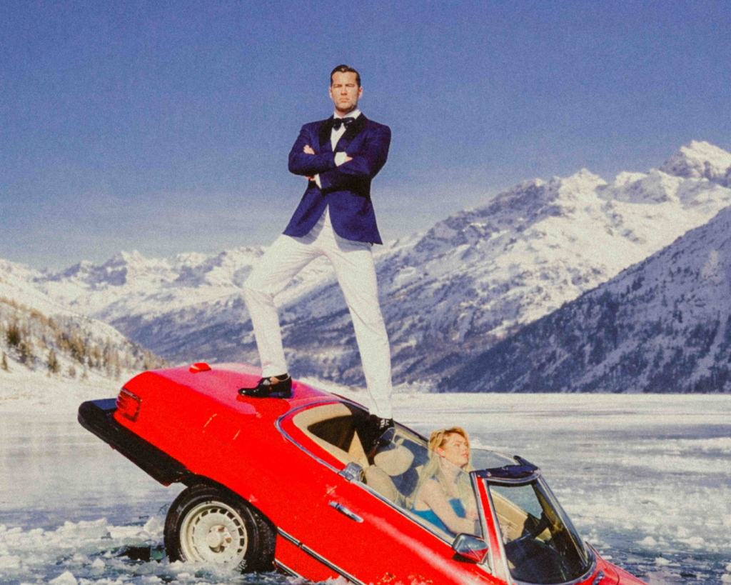 Man standing confidently on top of a red sports car stuck in icy water, with snowy mountains in the background.